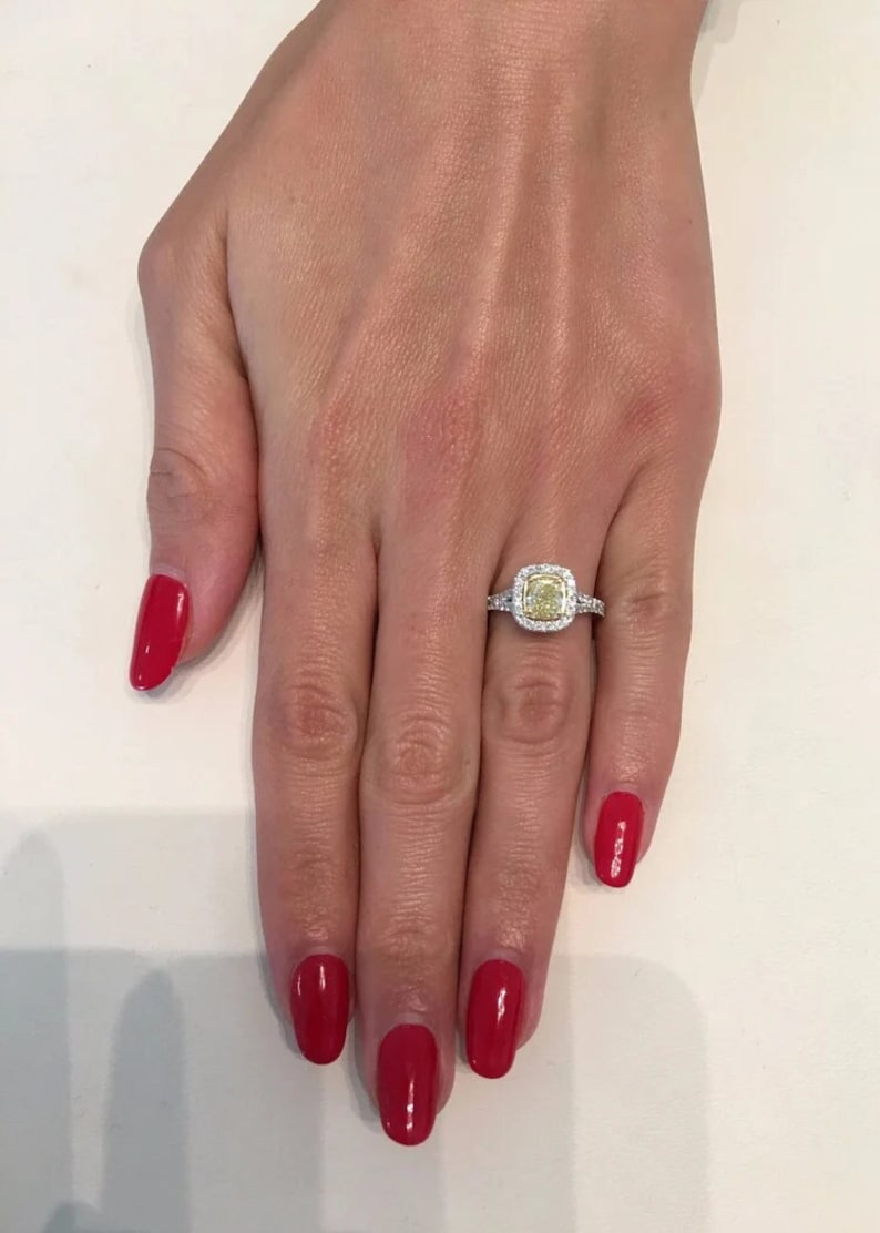 May include: A white gold engagement ring with a cushion-cut yellow diamond center stone surrounded by a halo of smaller diamonds. The ring is on a woman's finger with red nail polish.