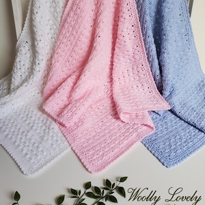 Op de afbeelding: Drie handgemaakte gehaakte dekens in wit, roze en lichtblauw. De dekens hebben ingewikkelde open patronen en zijn over een wit oppervlak gedrapeerd. De dekens zijn zacht en knus, perfect voor een baby of kind.