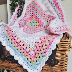May include: A crocheted baby blanket and hat set in white, with a colourful granny square pattern in pastel shades of pink, yellow, and blue. The blanket has a scalloped edge and is displayed on a wicker basket. The text "Woolly Lovely" is visible in the bottom right corner.