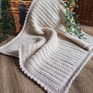 May include: A cream-colored, crocheted blanket with a scalloped edge. The blanket has a textured, ribbed pattern and a subtle gradient of light purple along the border. The text "Woolly Lovely" is visible in the bottom left corner. The blanket is folded on a wooden surface.