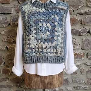 May include: A handmade, blue and grey crochet vest with a square pattern, layered over a white collared shirt. The vest is displayed on a wooden mannequin against a brick wall. The text "Woolly Lovely" is at the bottom.