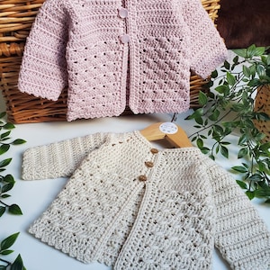 May include: Two handmade crochet baby cardigans. One is a soft pink with three pink buttons, the other is cream-coloured with three wooden buttons. Both have a textured stitch pattern and are displayed on wooden hangers.
