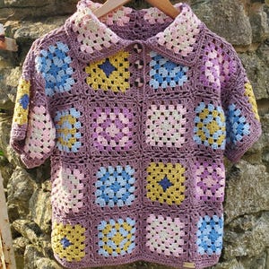 Puede incluir: Un cárdigan de ganchillo con cuello y mangas cortas. El cárdigan está hecho de cuadrados de la abuela en tonos de rosa, azul, amarillo y marrón.