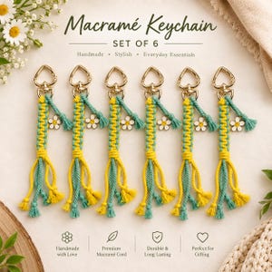 Puede incluir: Un conjunto de seis llaveros de macramé con cordones trenzados amarillos y verde azulado, cierres de mosquetón dorados en forma de corazón y pequeños dijes de margaritas blancas. El texto Macramé Keychain SET OF 6 aparece en la imagen.