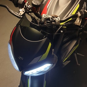 Może przedstawiać: Czarny motocykl z ż&oacute;łtymi i czerwonymi akcentami. Motocykl ma białe światło przednie i czarny zbiornik paliwa z napisem "Triumph".