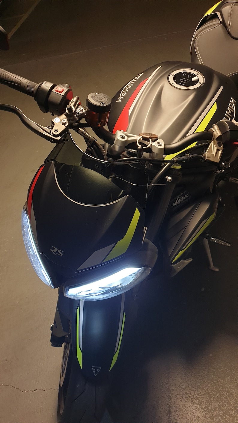 Może przedstawiać: Czarny motocykl z ż&oacute;łtymi i czerwonymi akcentami. Motocykl ma białe światło przednie i czarny zbiornik paliwa z napisem "Triumph".