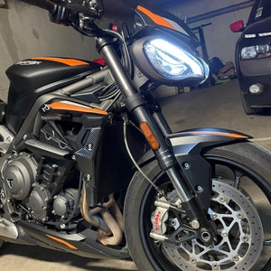 Może przedstawiać: Czarny motocykl z pomarańczowymi akcentami. Motocykl ma białe światło przednie i czarne przednie koło z srebrnym tarczą hamulcową. Motocykl jest zaparkowany w garażu.