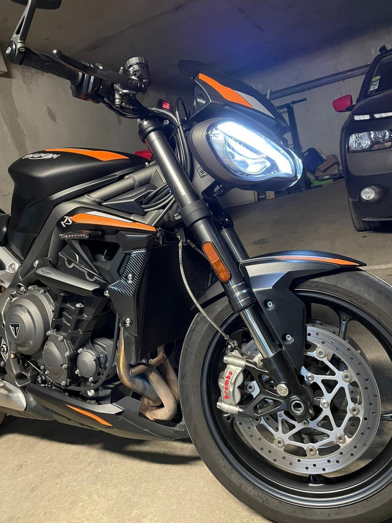 Może przedstawiać: Czarny motocykl z pomarańczowymi akcentami. Motocykl ma białe światło przednie i czarne przednie koło z srebrnym tarczą hamulcową. Motocykl jest zaparkowany w garażu.