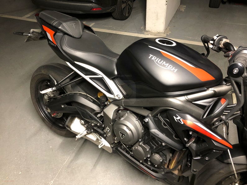 Może przedstawiać: Czarny motocykl Triumph z czerwonymi i białymi akcentami. Motocykl ma czarne sk&oacute;rzane siedzenie i czarny zbiornik paliwa z napisem "TRIUMPH" białymi literami.