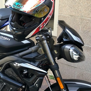 Może przedstawiać: Czarny motocykl Triumph z srebrną i czarną szybą, czarnym kaskiem z czerwonymi i białymi akcentami oraz napisem "TRIUMPH" na zbiorniku paliwa.