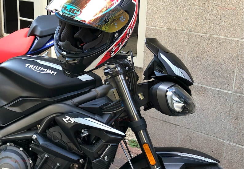 Może przedstawiać: Czarny motocykl Triumph z srebrną i czarną szybą, czarnym kaskiem z czerwonymi i białymi akcentami oraz napisem "TRIUMPH" na zbiorniku paliwa.