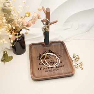 May include: A wooden tray with a mountain design and the text "The Brown Olivia & James 09.06.2024". A pearl necklace and a key holder with keys are on the tray.