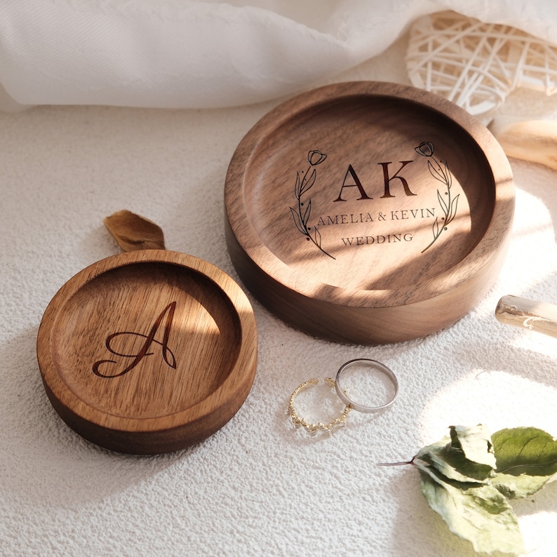 Puede incluir: Dos cajas de anillos de madera con iniciales grabadas y el texto "Amelia & Kevin Wedding". La caja m&aacute;s grande tiene un dise&ntilde;o floral y la caja m&aacute;s peque&ntilde;a tiene una sola inicial.