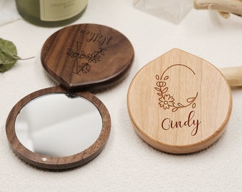 Puede incluir: Dos espejos compactos de madera con diseños florales grabados y nombres. El espejo de la izquierda está cerrado y tiene un acabado de madera oscura. El espejo de la derecha está abierto y tiene un acabado de madera clara. Los nombres "Allie" y "Cindy" están grabados en los espejos.