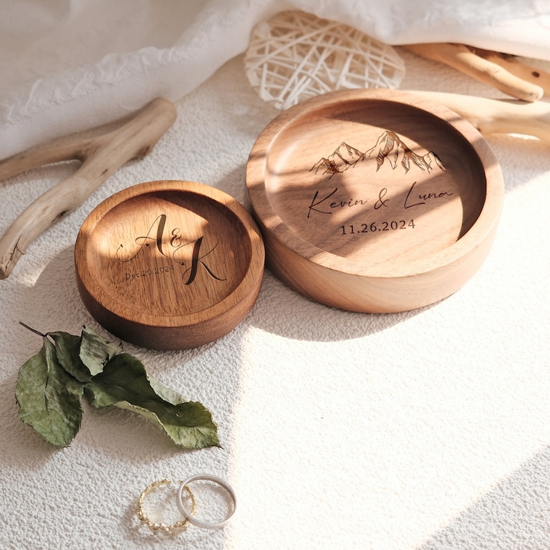 Puede incluir: Dos cajas de anillos de madera con dise&ntilde;os grabados. La caja m&aacute;s grande tiene un dise&ntilde;o de cordillera y el texto "Kevin & Luna 11.26.2024". La caja m&aacute;s peque&ntilde;a tiene las iniciales "A & K" y la fecha "Dec 20, 2024".