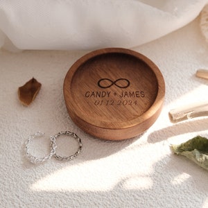Puede incluir: Una caja de anillos de madera con un s&iacute;mbolo de infinito y el texto "CANDY + JAMES 01.12.2024" grabado en la parte superior. Dos anillos de plata se colocan sobre una superficie blanca junto a la caja.