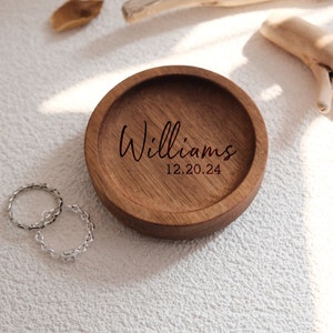 Puede incluir: Un plato de madera para anillos con la inscripci&oacute;n "Williams 12.20.24" grabada en la parte superior. Dos anillos de plata se colocan junto al plato.