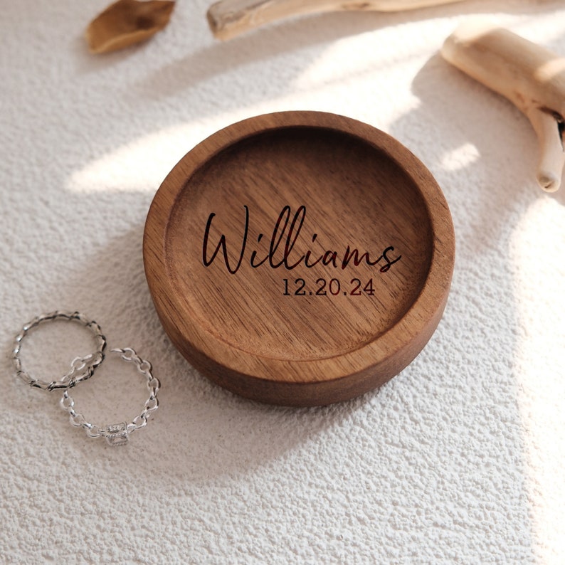 Puede incluir: Un plato de madera para anillos con la inscripci&oacute;n "Williams 12.20.24" grabada en la parte superior. Dos anillos de plata se colocan junto al plato.
