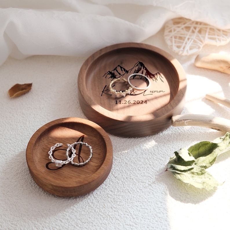 Puede incluir: Dos platos de anillos de madera, uno con un dise&ntilde;o de monta&ntilde;a y el texto "Kevin & Luna 11.26.2024" y el otro con la letra "A" grabada.