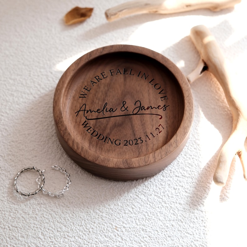 Puede incluir: Un plato de anillos redondo de madera con el texto "We are fall in love Amelia & James Wedding 2023.11.27" grabado en la parte superior.