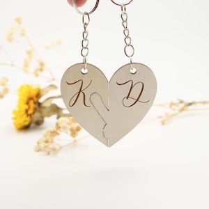 May include: Two wooden heart-shaped keychains with the letters "K" and "D" engraved on them. The keychains are connected by a silver chain.