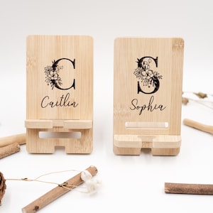 Può includere: Due supporti per telefono in bambù, ciascuno inciso con un'iniziale floreale e un nome. Uno presenta la lettera "C" con "Caitlin" sotto, e l'altro ha "S" con "Sophia". I supporti hanno una fessura per tenere un telefono.