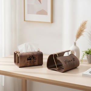 Puede incluir: Dos cajas de pañuelos de madera marrón oscuro con lados curvos y detalles de metal dorado. Una caja presenta un gráfico de montaña con el texto Kinda & Jack 07.08.2025, mientras que la otra muestra las iniciales S&K flanqueadas por hojas.
