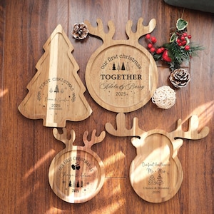 Pode incluir: Travessas de servir de Natal em madeira em forma de árvore de Natal e renas. Cada travessa é gravada com as palavras "Our First Christmas" e nomes, juntamente com o ano de 2025. As travessas são exibidas numa superfície de madeira.