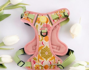 May include: A pink dog harness with a green and yellow floral pattern. The harness has a mesh lining and a black buckle. The harness has a yellow tag that says "Haus of Hound".