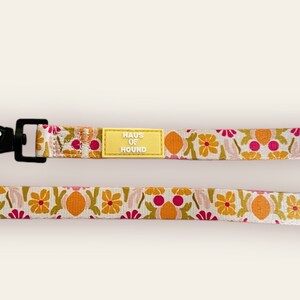May include: A white dog lead with a floral pattern in shades of pink, orange, and green. The lead has a black clip and a yellow tag that reads "HAUS OF HOUND".