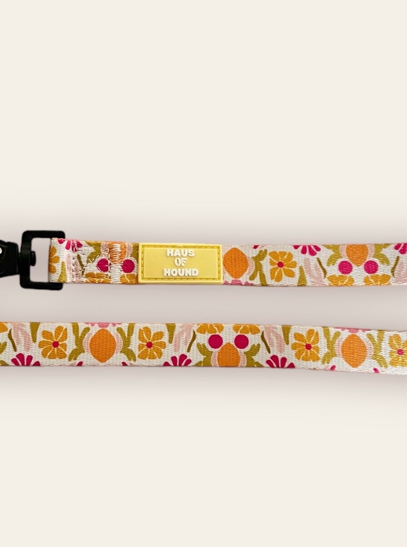 May include: A white dog lead with a floral pattern in shades of pink, orange, and green. The lead has a black clip and a yellow tag that reads "HAUS OF HOUND".