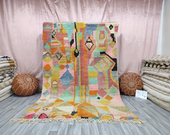 May include: A colorful Moroccan rug with geometric patterns in shades of pink, orange, yellow, blue, and green. The rug has a fringe border and is displayed on a light pink background.