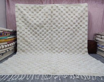 May include: A white, fluffy, hand-woven rug with a checkered pattern and fringe. The rug is made of natural fibers and has a soft, textured surface.