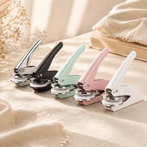 Pode incluir: Cinco carimbos de relevo manuais em metal dispostos em fila, com acabamentos em prateado, preto, verde menta, rosa claro e branco. Cada dispositivo possui uma alavanca longa, um mecanismo de dobradiça e uma placa circular na base.
