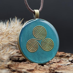 May include: A circular teal pendant featuring a gold-coloured triple spiral design, suspended from a brown cord. The pendant has a bronze-coloured metal border.
