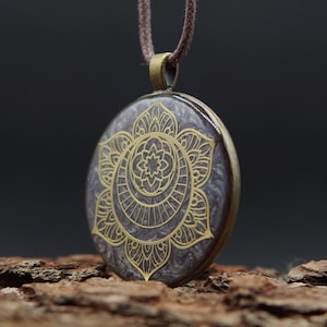 May include: A round pendant necklace featuring a dark gray, marbled resin base with a gold-colored metallic inlay of a symmetrical floral mandala design. The pendant is suspended from a brown cord and encased in a bronze-toned metal frame.