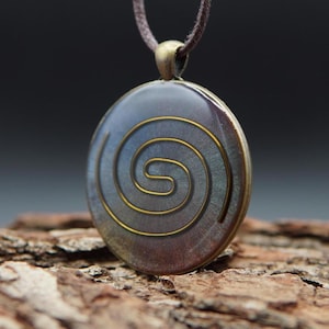 Op de afbeelding: Een ronde hangerketting met een gouden spiraalontwerp op een donkerblauwe achtergrond. De hanger is gemaakt van metaal en heeft een bruin leren koord.