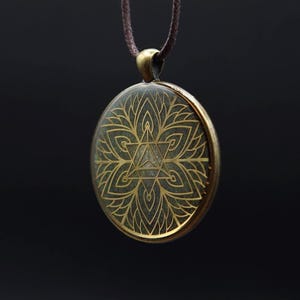 Könnte beinhalten: Ein runder Anhänger mit einem goldfarbenen Blumenmuster und einer sternförmigen Mitte. Der Anhänger hängt an einer braunen Kordel. Das filigrane Muster hebt sich vor einem dunklen Hintergrund ab und betont die detaillierte Handwerkskunst des Schmucks.