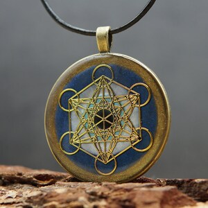 Könnte beinhalten: Ein runder Anhänger mit einem Metatron's Cube-Design. Der Anhänger hat ein goldfarbenes geometrisches Muster auf einem blau-weißen Hintergrund mit einem schwarzen Zentrum. Er hängt an einer schwarzen Kordel.