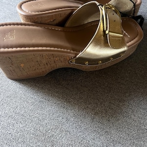 Puede incluir: Sandalias doradas con cu&ntilde;a, suelas estilo corcho y hebillas doradas. Las sandalias tienen un acabado dorado met&aacute;lico en las correas y est&aacute;n tachonadas con peque&ntilde;os remaches dorados. Las suelas son de color marr&oacute;n claro.