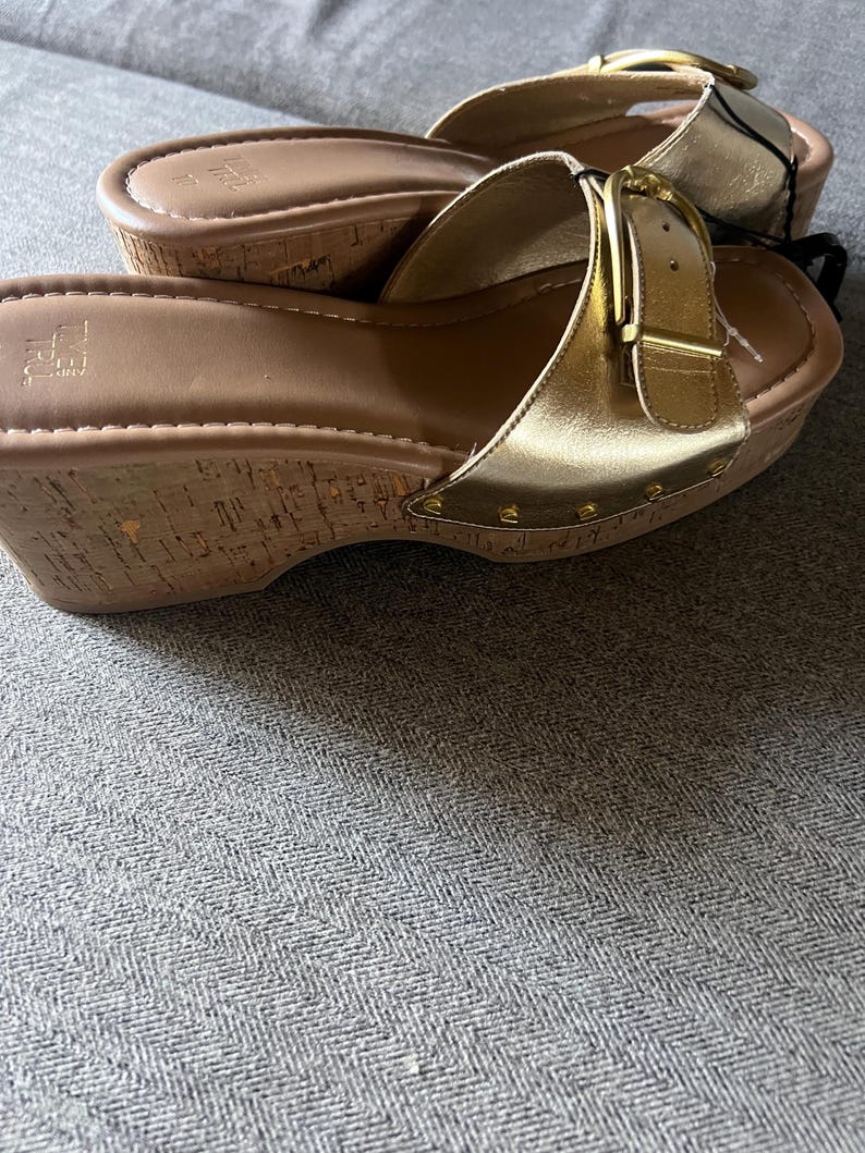 Puede incluir: Sandalias doradas con cu&ntilde;a, suelas estilo corcho y hebillas doradas. Las sandalias tienen un acabado dorado met&aacute;lico en las correas y est&aacute;n tachonadas con peque&ntilde;os remaches dorados. Las suelas son de color marr&oacute;n claro.