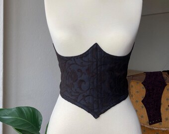 May include: A black corset with a floral pattern on a white mannequin. The corset has a pointed bottom and a low neckline.