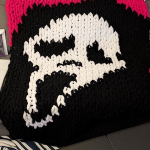 Può includere: Una coperta a maglia grossa con un motivo a teschio. La coperta è principalmente nera, con un grande teschio bianco al centro. Il bordo superiore è rosa acceso, creando un contrasto sorprendente. Ideale per aggiungere un tocco unico a qualsiasi stanza.