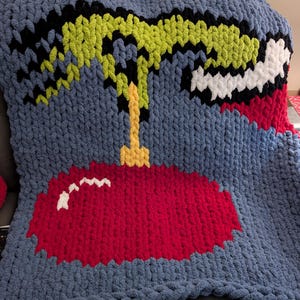 May include: A handmade, chunky knit blanket in shades of blue, red, black, white, and yellow. The blanket features a large, stylized Christmas ornament design. The ornament is red with white accents, and the top of the ornament is yellow.