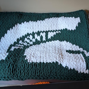 May include: A chunky knit blanket in dark green with a large white design resembling a map of Michigan. The blanket is rectangular and appears soft and cozy, suitable for a couch or bed. The design is prominently displayed against the green background.