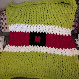 May include: A large, hand-knitted blanket in a festive design. The blanket features a green base with white and red stripes, and a black square in the center. The chunky knit adds texture and warmth to this cozy home decor item.