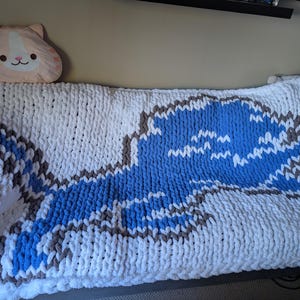 Könnte beinhalten: Eine grob gestrickte Decke in Weiß mit einem großen blauen und grauen Design. Ein katzenförmiges Kissen in Pfirsich- und Weißtönen liegt links auf der Decke. Die Decke wirkt dick und weich.