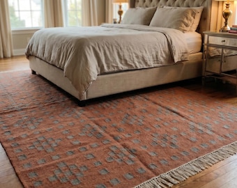 May include: A bedroom with a tufted headboard bed, beige bedding, and a patterned rug in shades of orange and gray. Two bedside tables with lamps flank the bed. The room has large windows with cream-colored curtains.