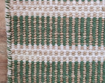 May include: Close-up of a rectangular rug with a striped pattern. The rug features alternating bands of green, off-white, and tan colors. The edges are finished with a green border.