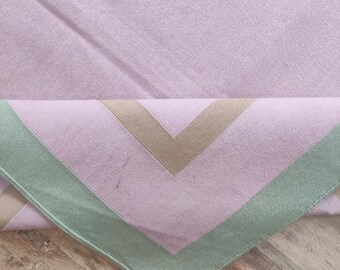 May include: A folded, light purple textile with a geometric design. The design features diagonal tan stripes and a green border. The textile is resting on a wooden surface.